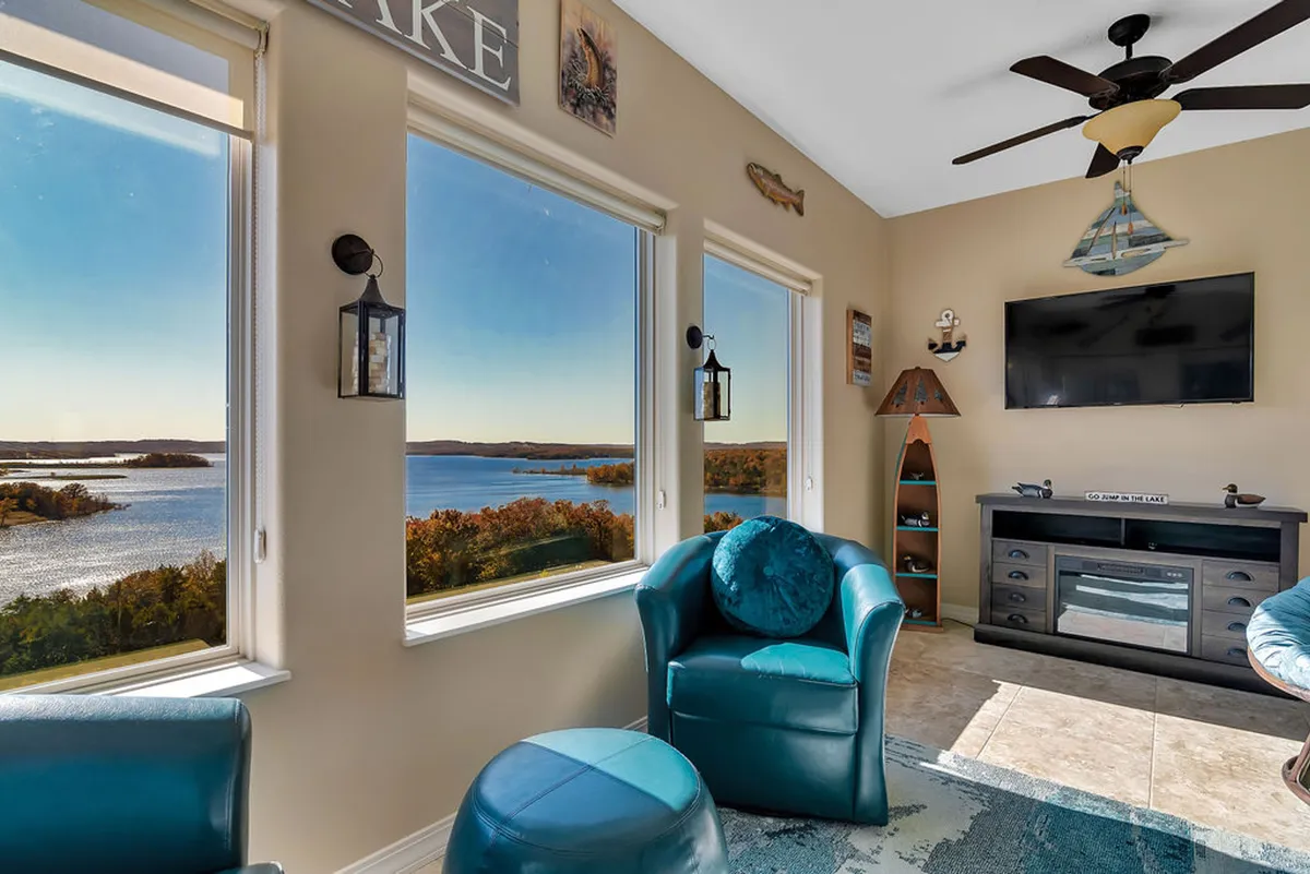 A cozy area featuring large windows with serene lake views, comfortable teal chairs, and a TV unit. The natural light and serene mood create an ideal retreat for relaxation, reading, or socializing.