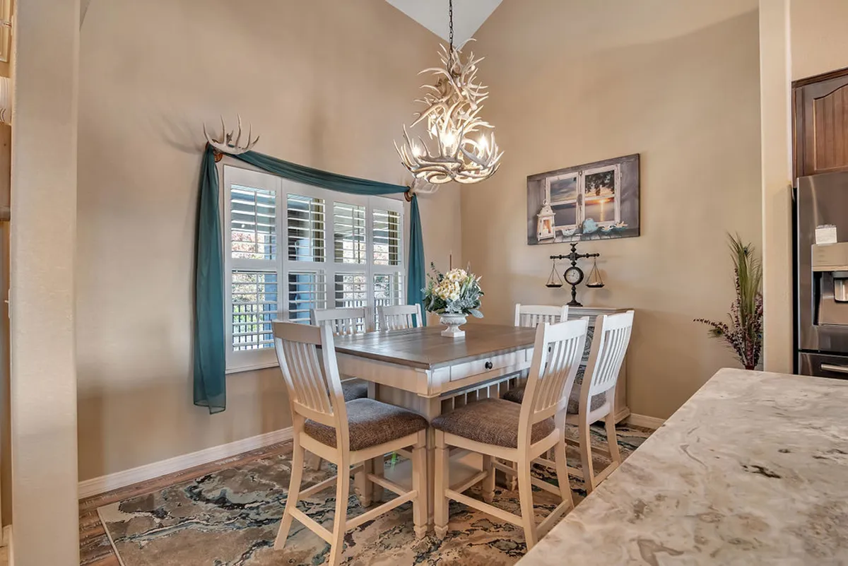 A cozy dining area with a welcoming table set beneath a chandelier, sunlight pouring in through window shutters, and decorative artwork on the wall. The setting evokes warmth and togetherness, suggesting celebrations, shared meals, and festive gatherings.