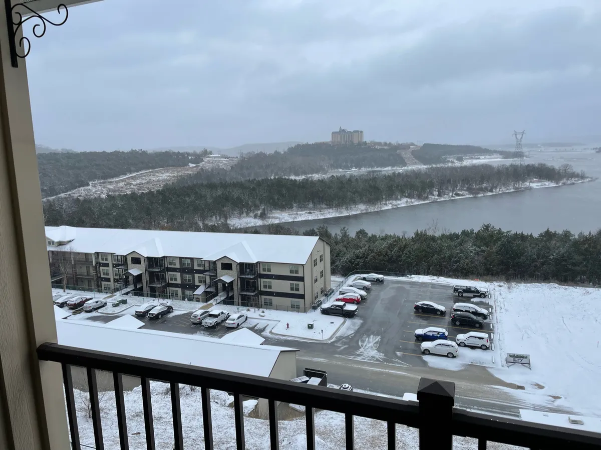 A serene winter landscape with a snow-covered river, an apartment complex in the background, and power lines stretching across the horizon. The scene is tranquil and peaceful, offering a sense of close proximity to nature from the comfort of a residential setting.