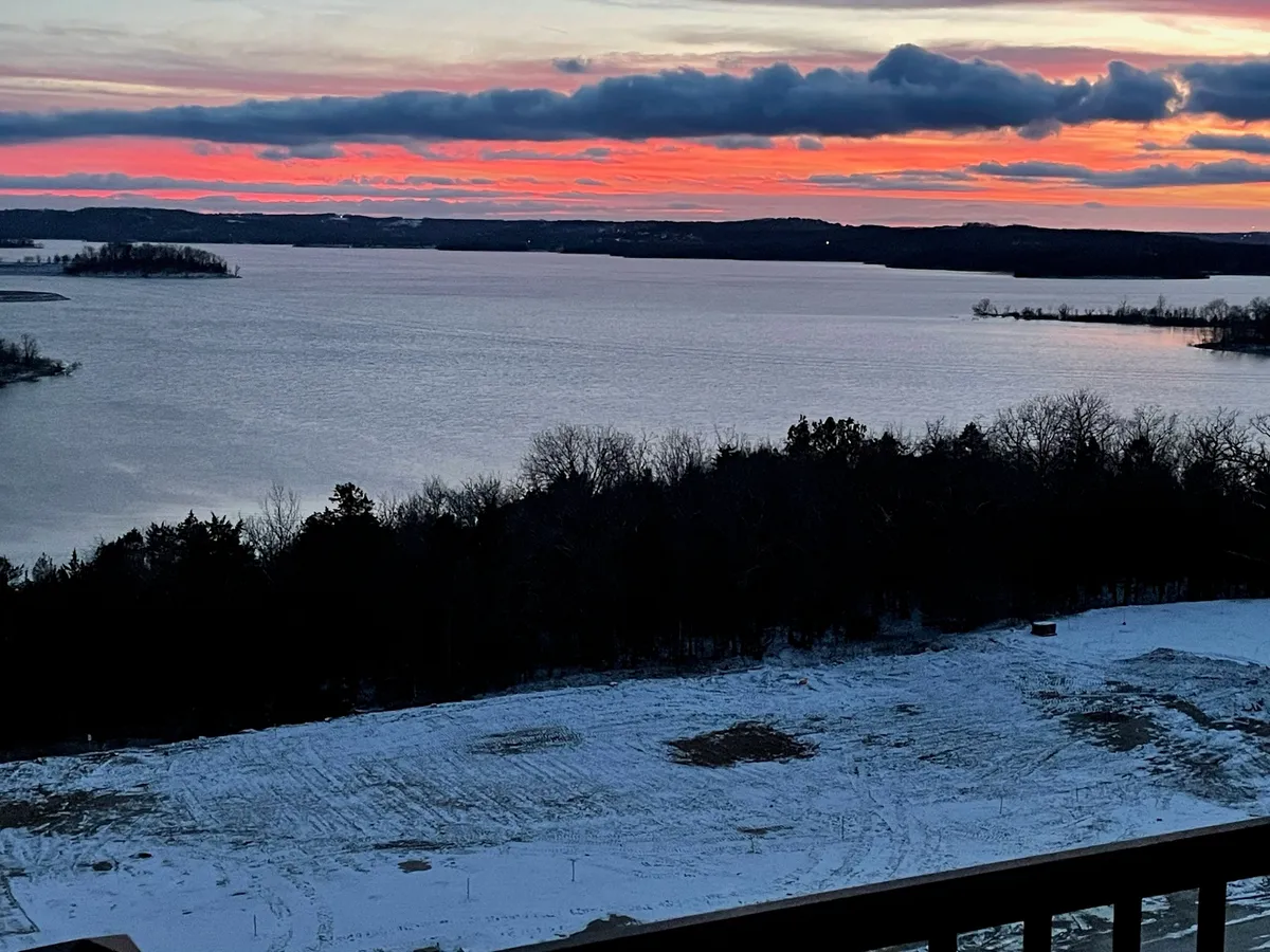This image showcases a breathtaking sunset over the lake, with the tranquil water bordered by a forest line and snow-covered ground in the foreground. The peaceful setting invites viewers to imagine relaxing by the water during golden hour.