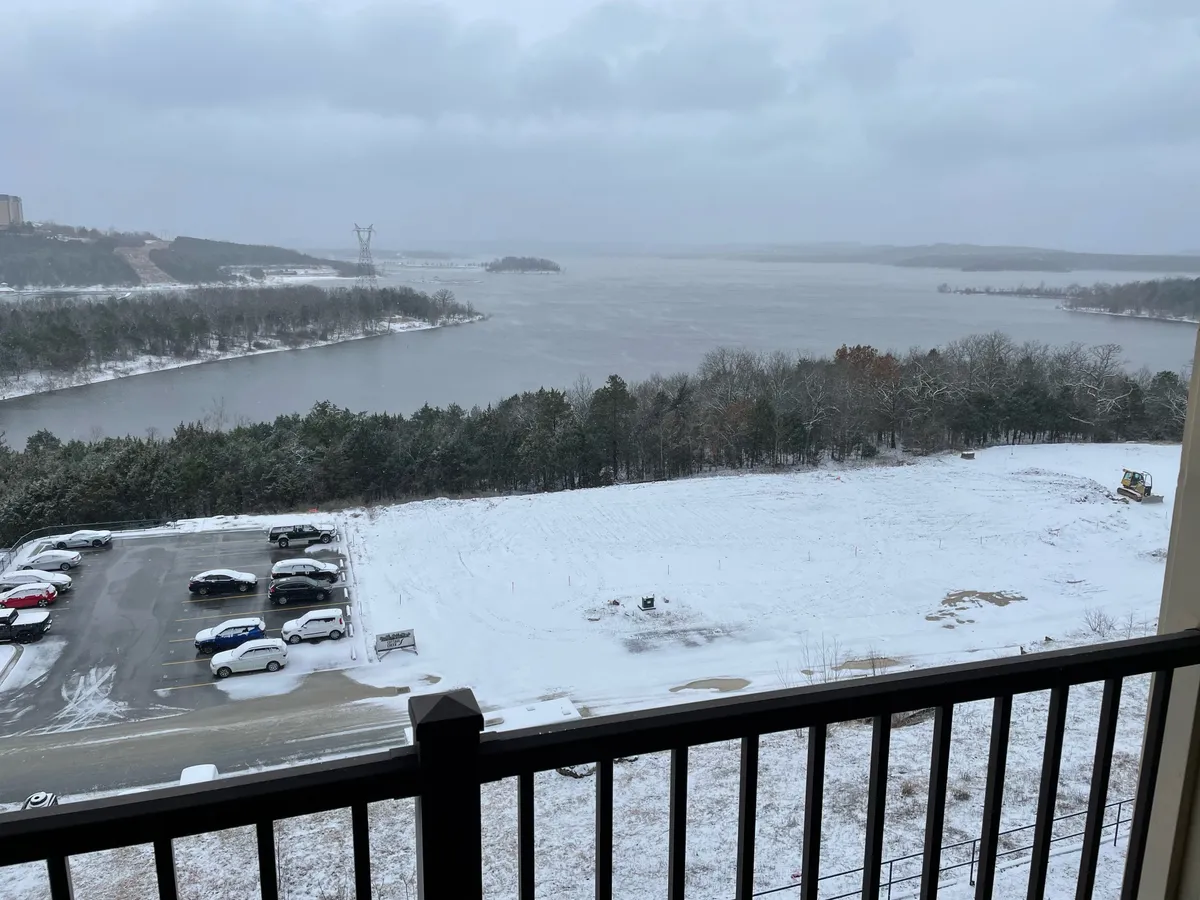A panoramic view of a snow-blanketed lake, bordered by distant trees and a cloudy sky. The image includes a glimpse of a parking lot, emphasizing easy lake access from the property.
