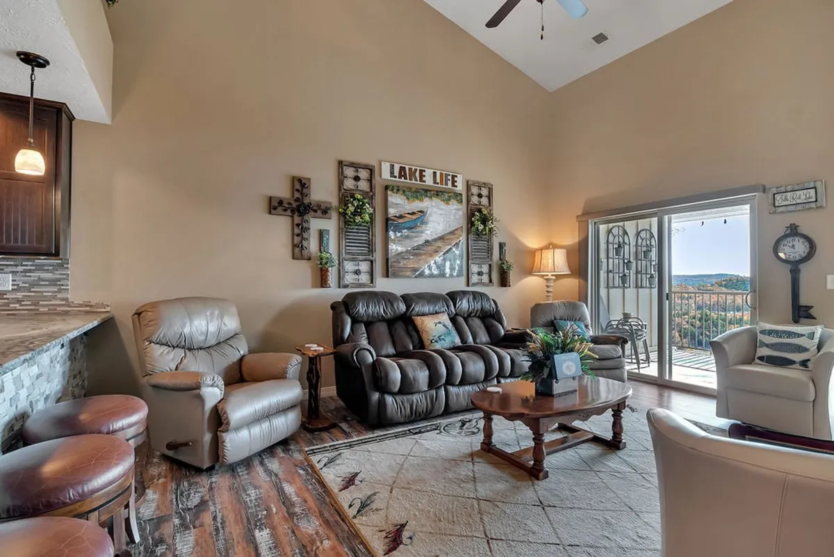 A bright, inviting living area with large windows that frame scenic outdoor views. Reclining chairs and a coffee table create a cozy space for relaxing or entertaining, while the lake-themed wall art connects the interior to the surrounding nature.