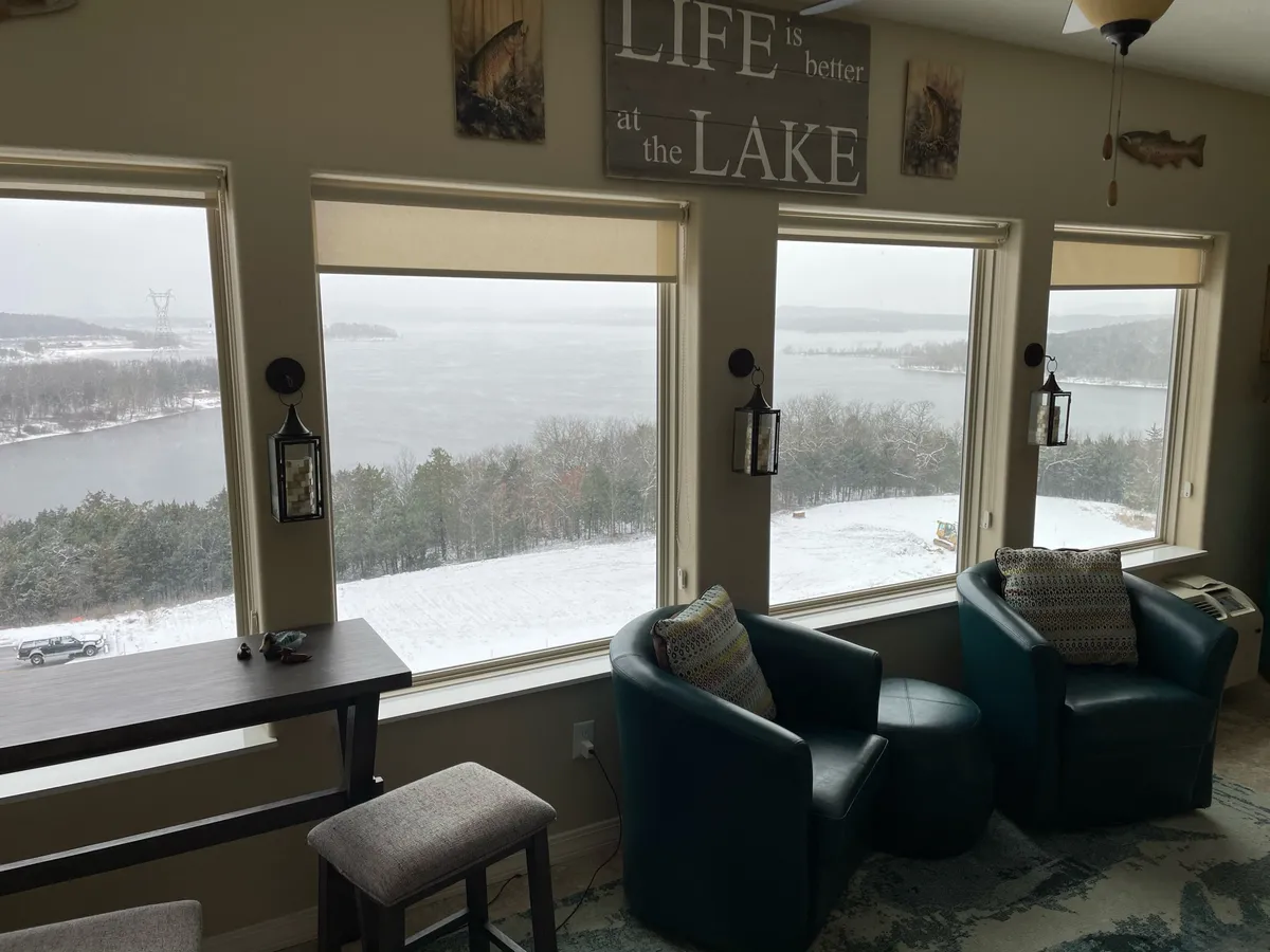 A sunroom with large windows framing sweeping lake views, comfortable seating, decorative touches, and hanging lanterns. The arrangement creates an inviting space that encourages relaxation, reading, and gathering with friends or family while enjoying the scenic backdrop.
