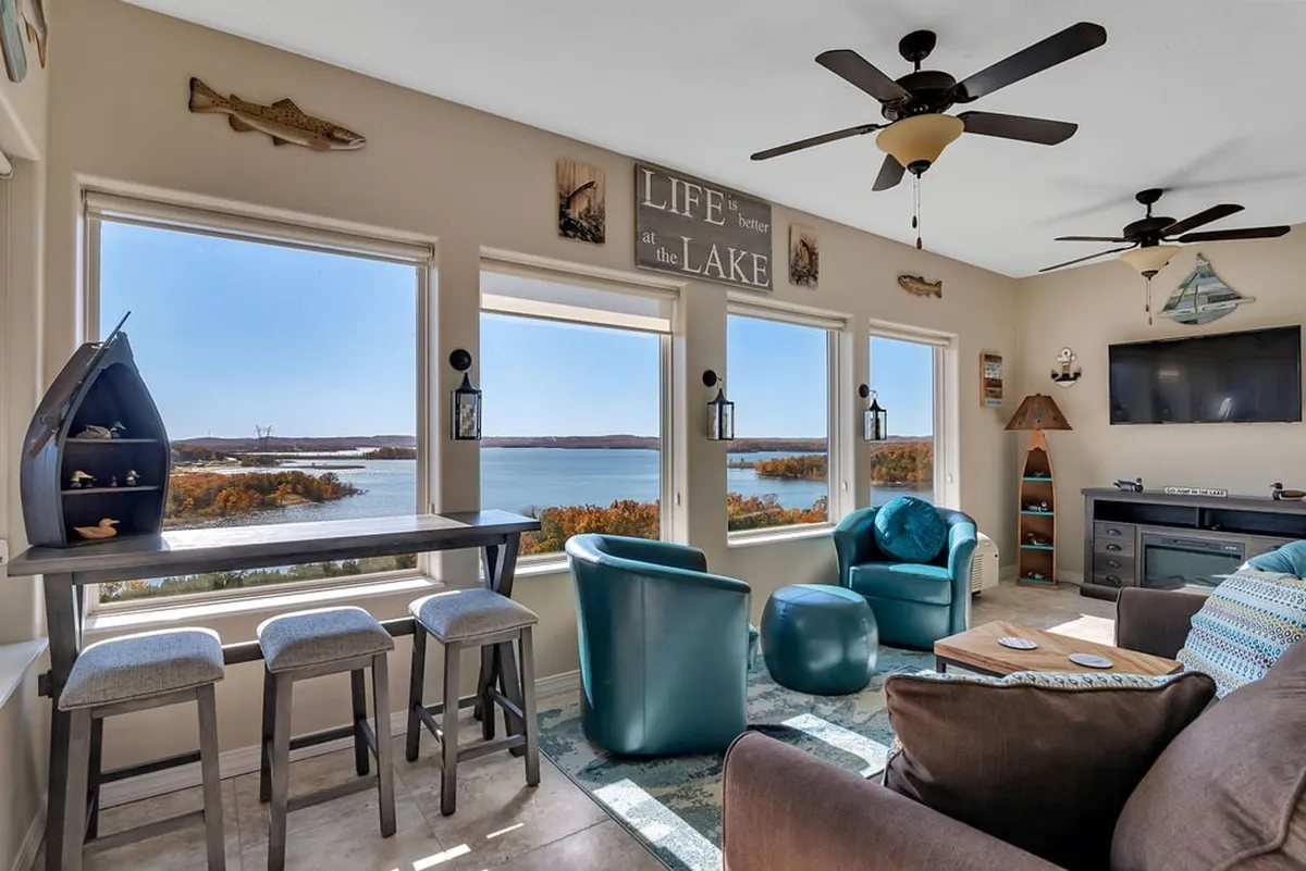 The image showcases a bright and spacious penthouse living area with expansive windows framing a stunning view of Table Rock Lake. Plush, comfortable seating invites guests to relax, while decorative wall signs add a warm, personal touch. The setting bathes in natural light, creating a serene, welcoming mood perfect for family gatherings or quiet moments enjoying the scenery.